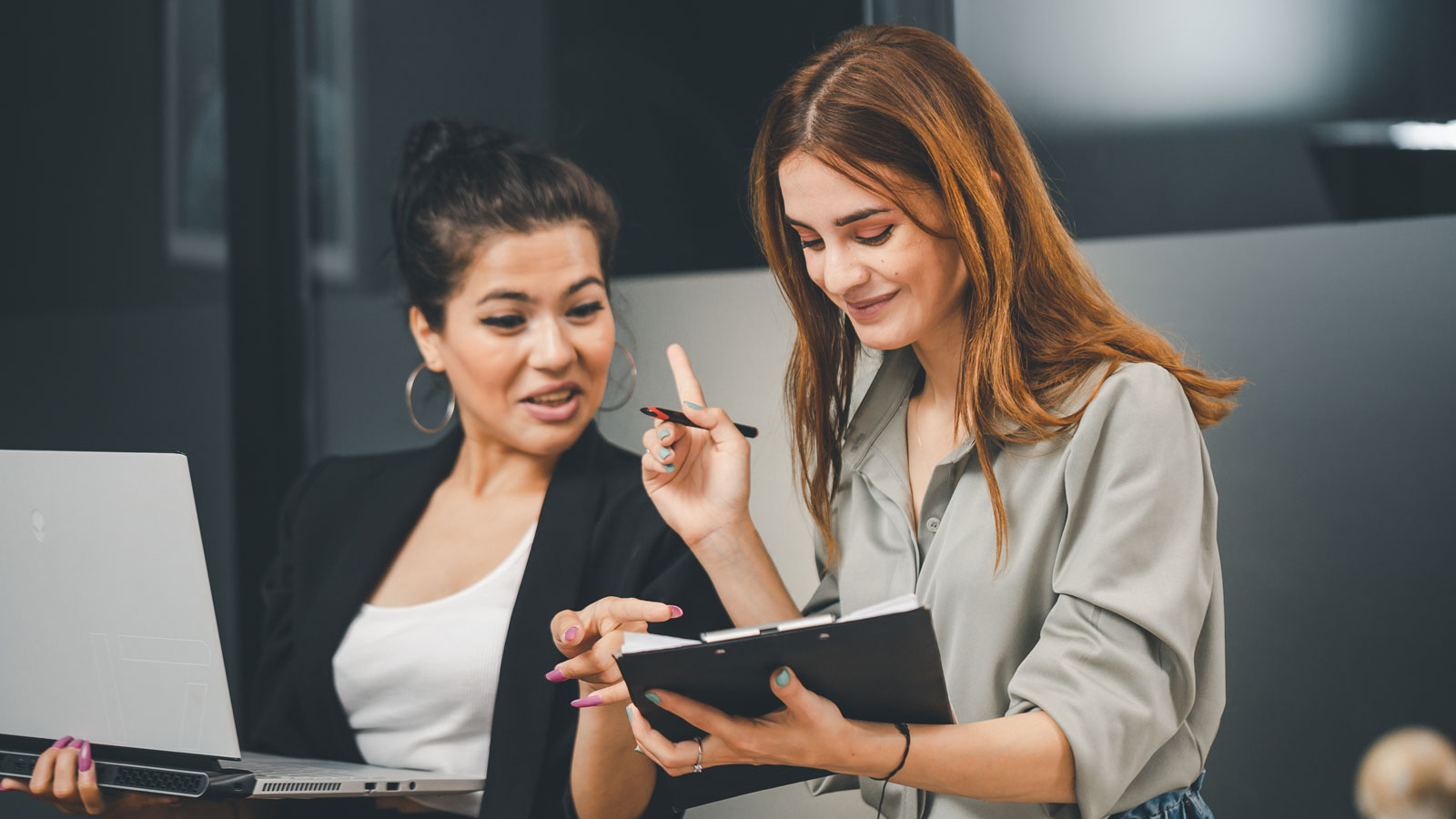 Zwei Frauen im Gespräch, eine hält einen Laptop, die andere zeigt mit einem Stift auf ein Tablet